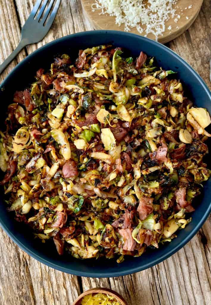 Large black bowl with cooked shredded Brussels sprouts, pieces of prosciutto, and seasonings. There is a black fork, a small wood dish with lemon zest, and a small round wood board with grated white cheese. Items are on a wood surface.