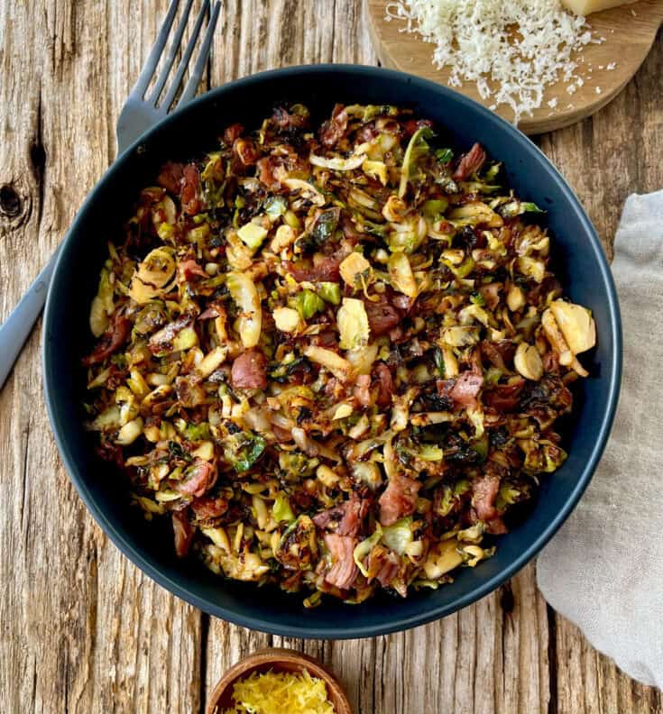 Large black bowl with cooked shredded Brussels sprouts, pieces of prosciutto, and seasonings. There is a black fork, a small wood dish with lemon zest, a small round wood board with grated white cheese, and a light brown cloth napkin. Items are on a wood surface.