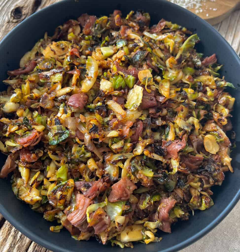 Large black bowl with cooked shredded Brussels sprouts, pieces of prosciutto, and seasonings. Bowl is on a wood surface.