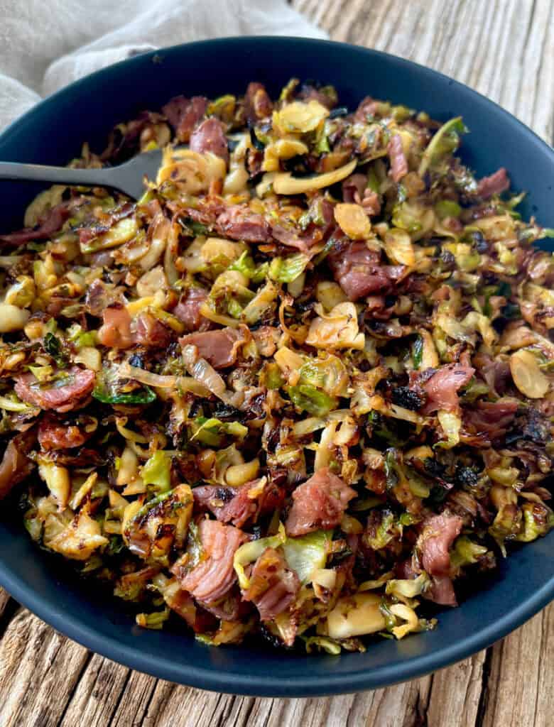 Large black bowl with cooked shredded Brussels sprouts, pieces of prosciutto, and seasonings. There is a black fork resting in the bowl. There is a light brown cloth napkin laying in the background. Items are on a wood surface.