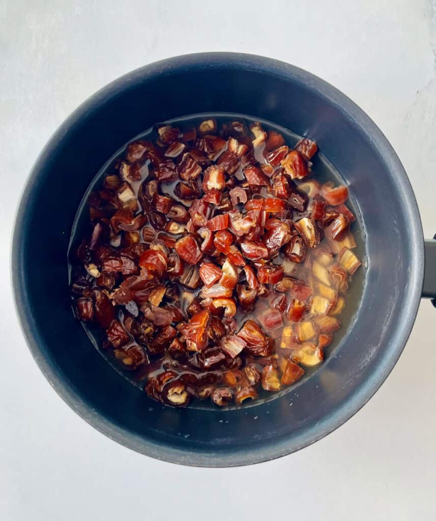 Dark saucepan with chopped dates, water, syrup, and vanilla in it. Pan is on a white surface.