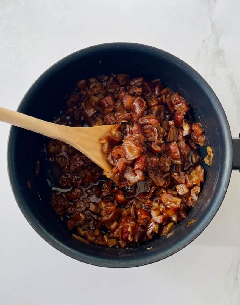Dark saucepan with chopped dates, water, syrup, and vanilla in it. There is a wooden spoon resting in the pan. Pan is on a white surface.
