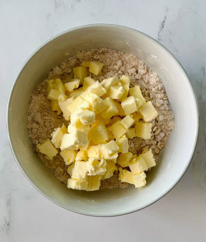 White bowl with an oatmeal mixture and butter cubes in it. Bowl is on a white surface.