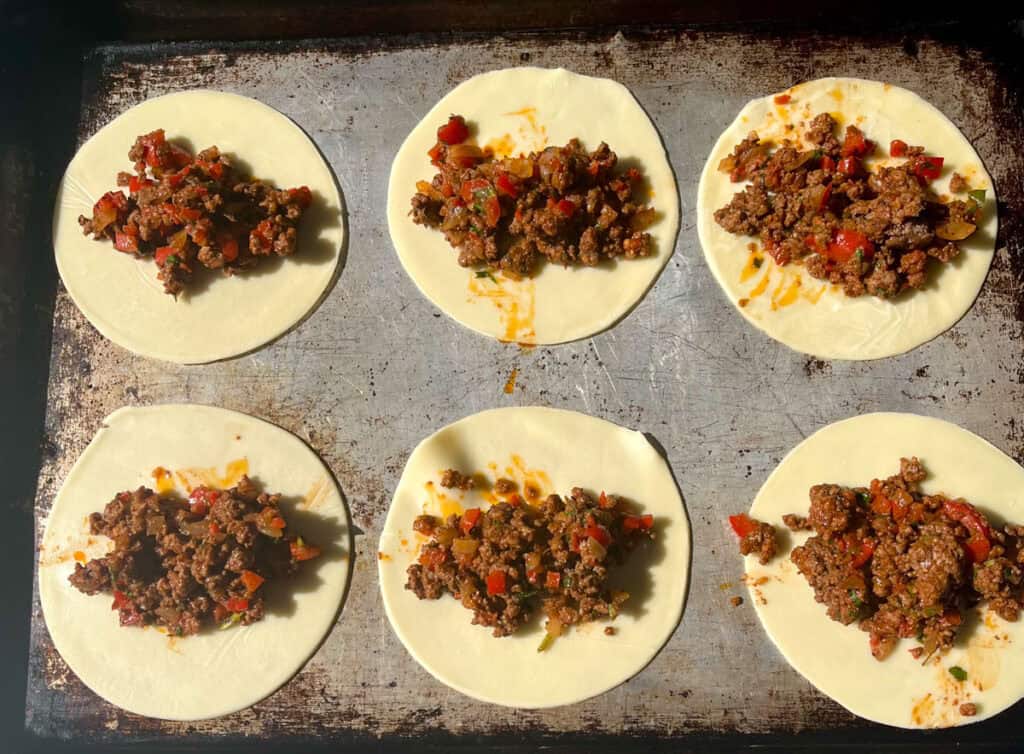Six empanada dough discs with meat filling on top.