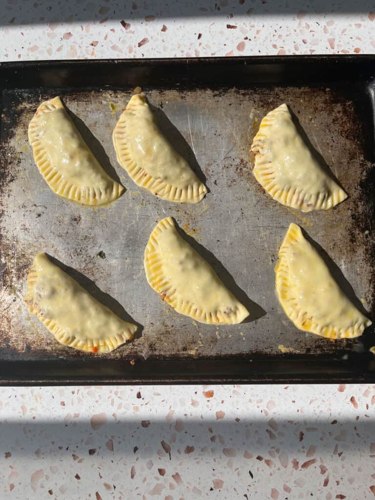 Six unbaked empanadas on a baking sheet.