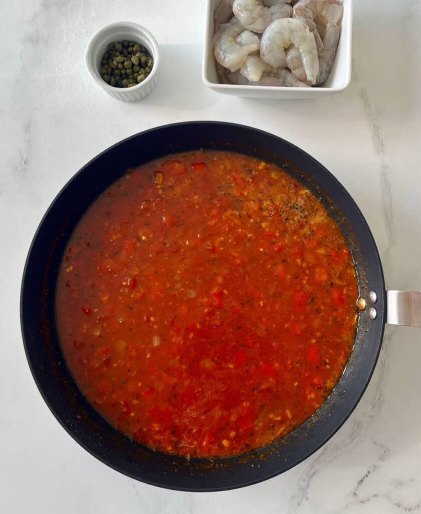 A pan filled with tomato sauce, a bowl of raw shrimp and a bowl of capers.