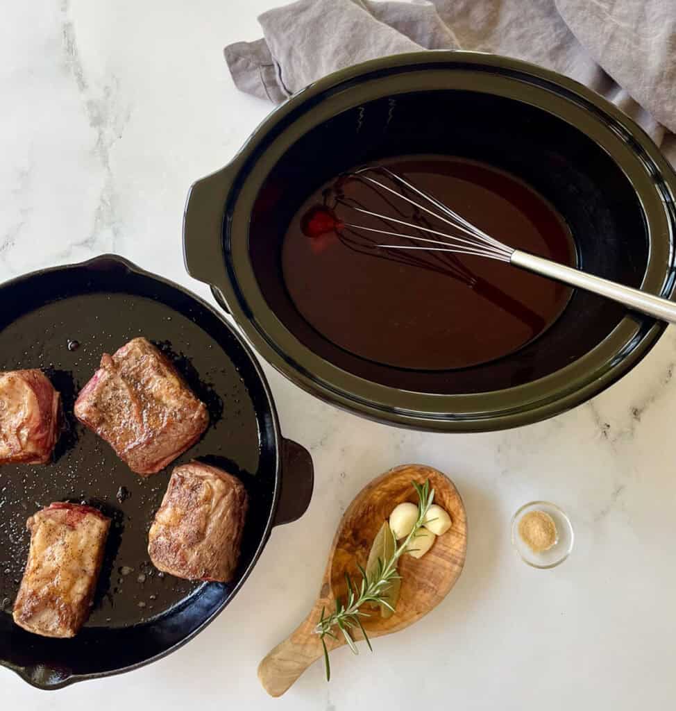 A slow cooker with sauce, a skillet with meat and herbs on a marble counter.