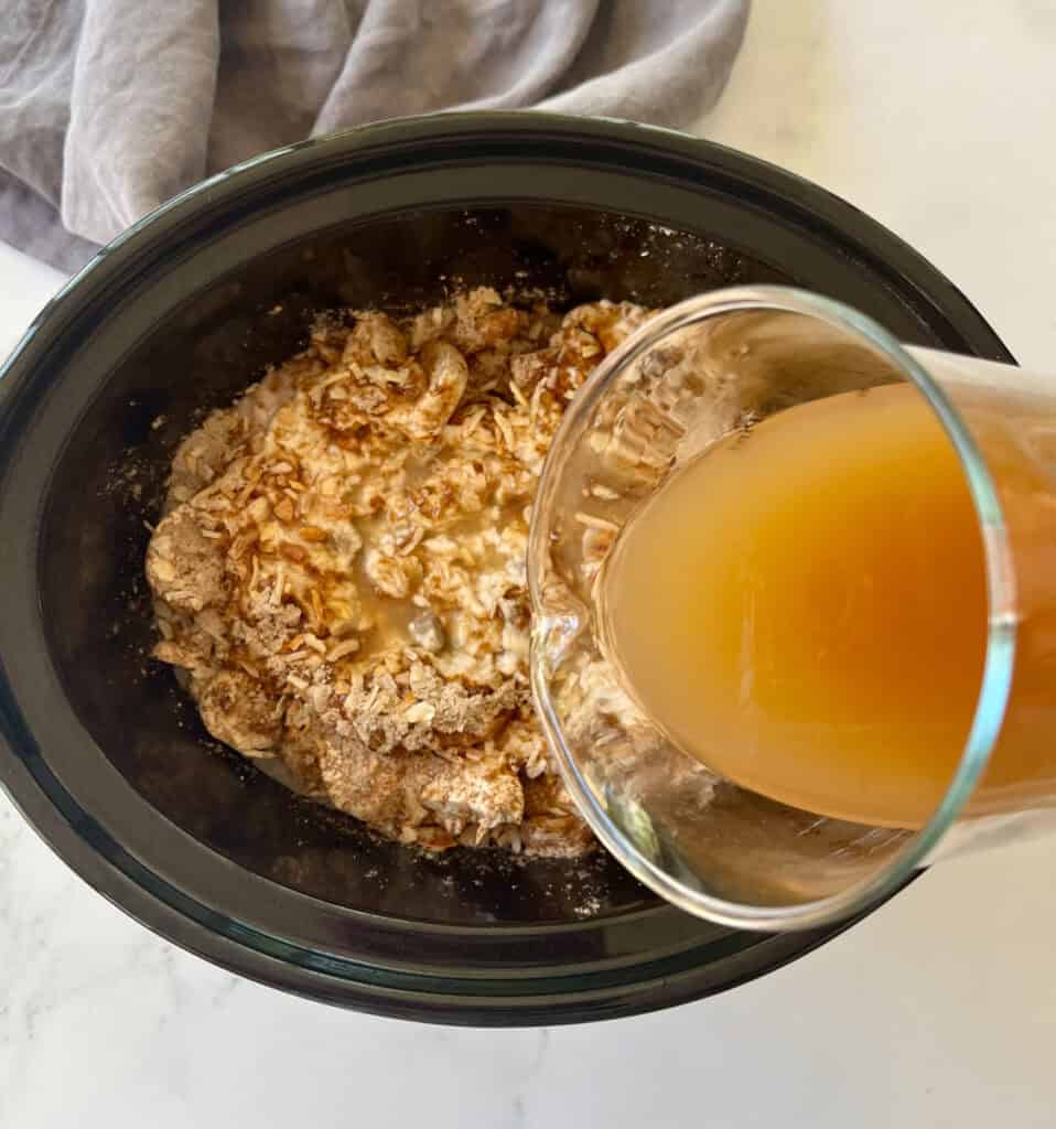 A hand pouring a glass of chicken broth into a slow cooker.
