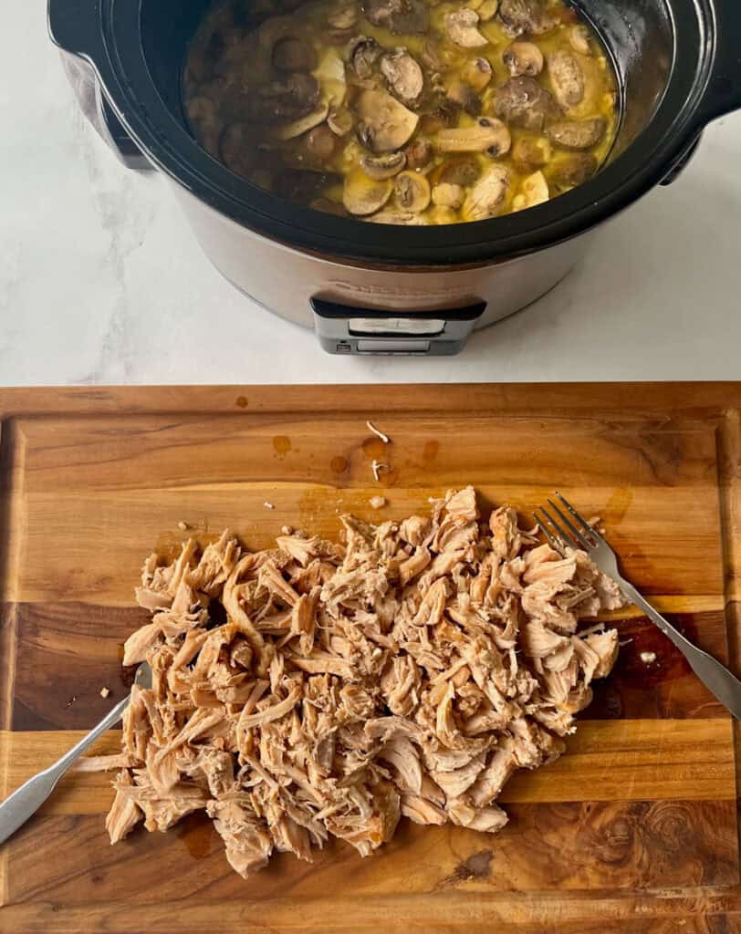 Shredded pork chops on a wooden board and mushroom soup in slow cooker.
