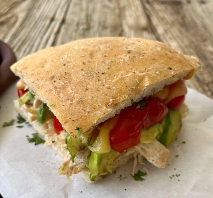 A chicken sandwich with tomatoes, bacon, and avocado on a piece of paper.