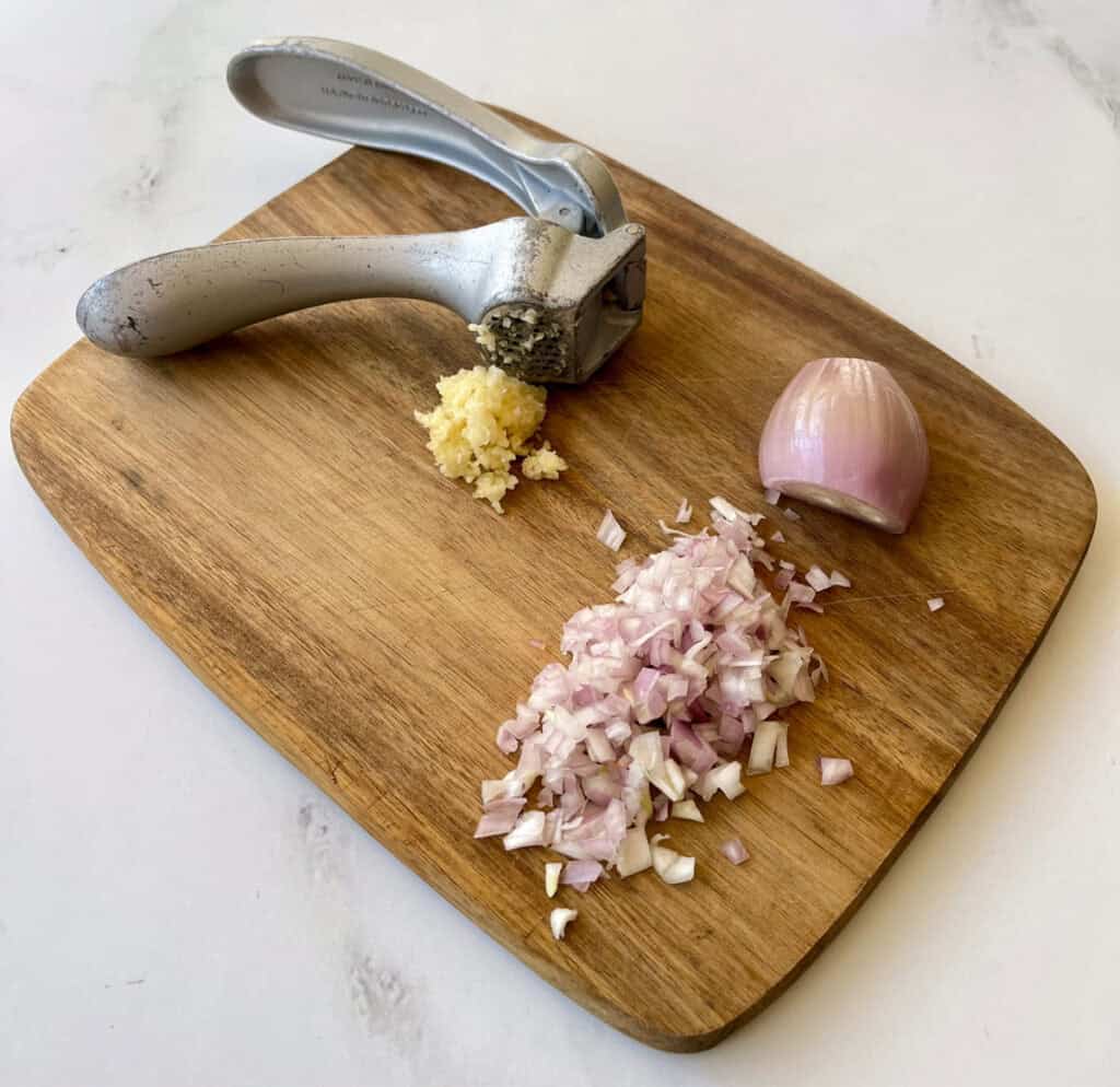 A cutting board with garlic, onion and a garlic presser.