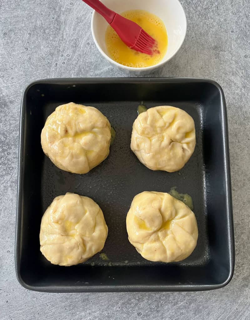 Four unbaked apple dumplings with an egg wash in a baking pan.