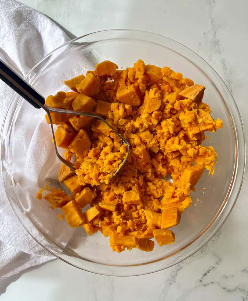 A bowl filled with cubed sweet potatoes. There is a masher utensil resting in the bowl.