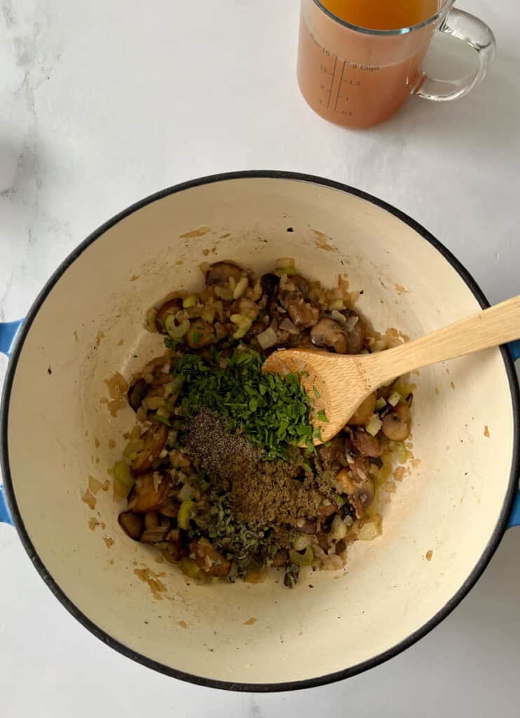 Sautéed mushrooms and herbs in a cooking pot with a wooden spoon and broth in the background.