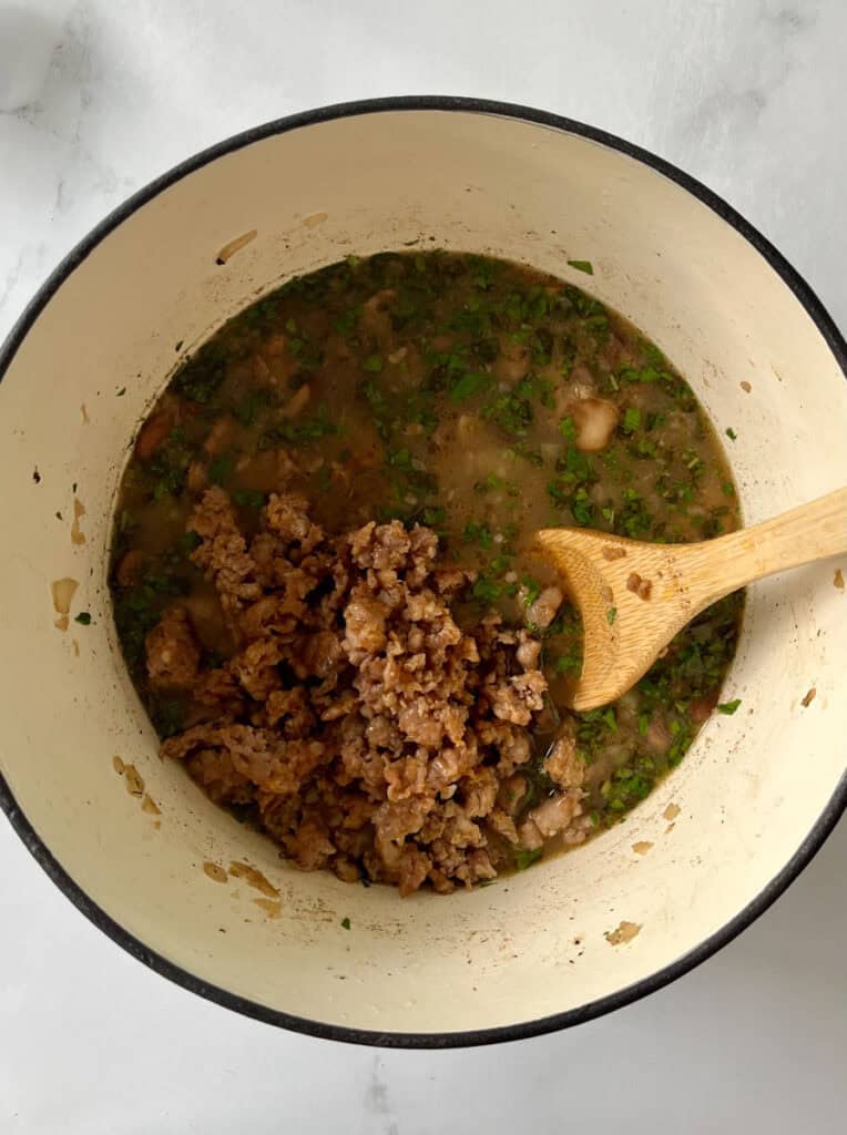 Cooked crumbled sausage in a pot with herbs, vegetables, and chicken broth in a large pot on a white counter.