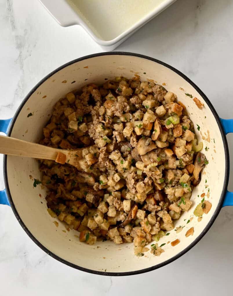 Cooked sausage with cubed bread, vegetables, and herbs in a white pot on a kitchen countertop.