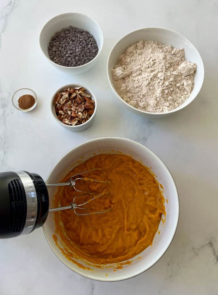 Pumpkin mixture being whipped with hand mixer in a white bowl, with baking ingredients nearby.