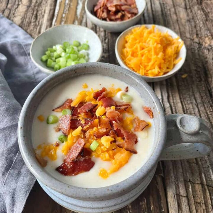 Creamy potato soup topped with bacon, cheese, and green onions on rustic wooden table.