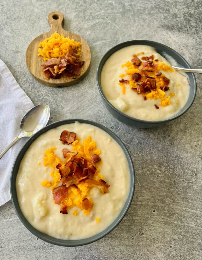Two bowls of creamy potato soup with crispy bacon and shredded cheese on a gray surface.