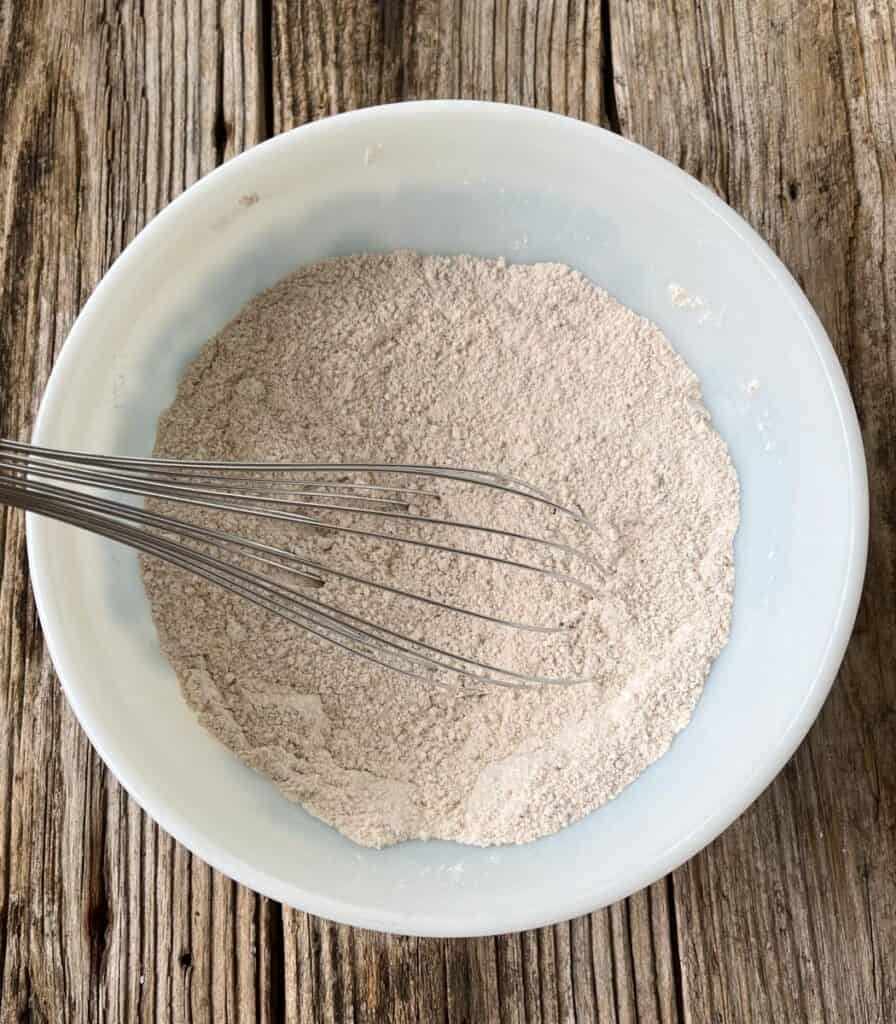 Large mixing bowl with a flour mixture and a whisk in it on a wooden surface.