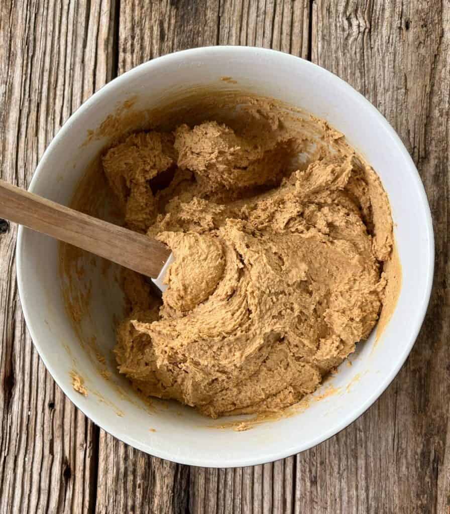 Large mixing bowl with cookie dough and a rubber spatula in it on a wooden surface.