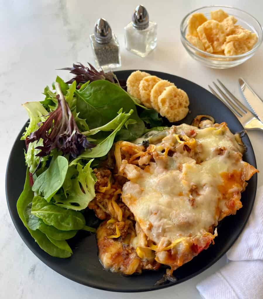 Zucchini sausage casserole served with fresh greens and crispy parmesan cheese crisps on a black plate.