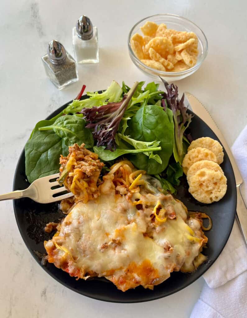 Zucchini sausage casserole served with fresh greens and crispy parmesan cheese crisps on a black plate.