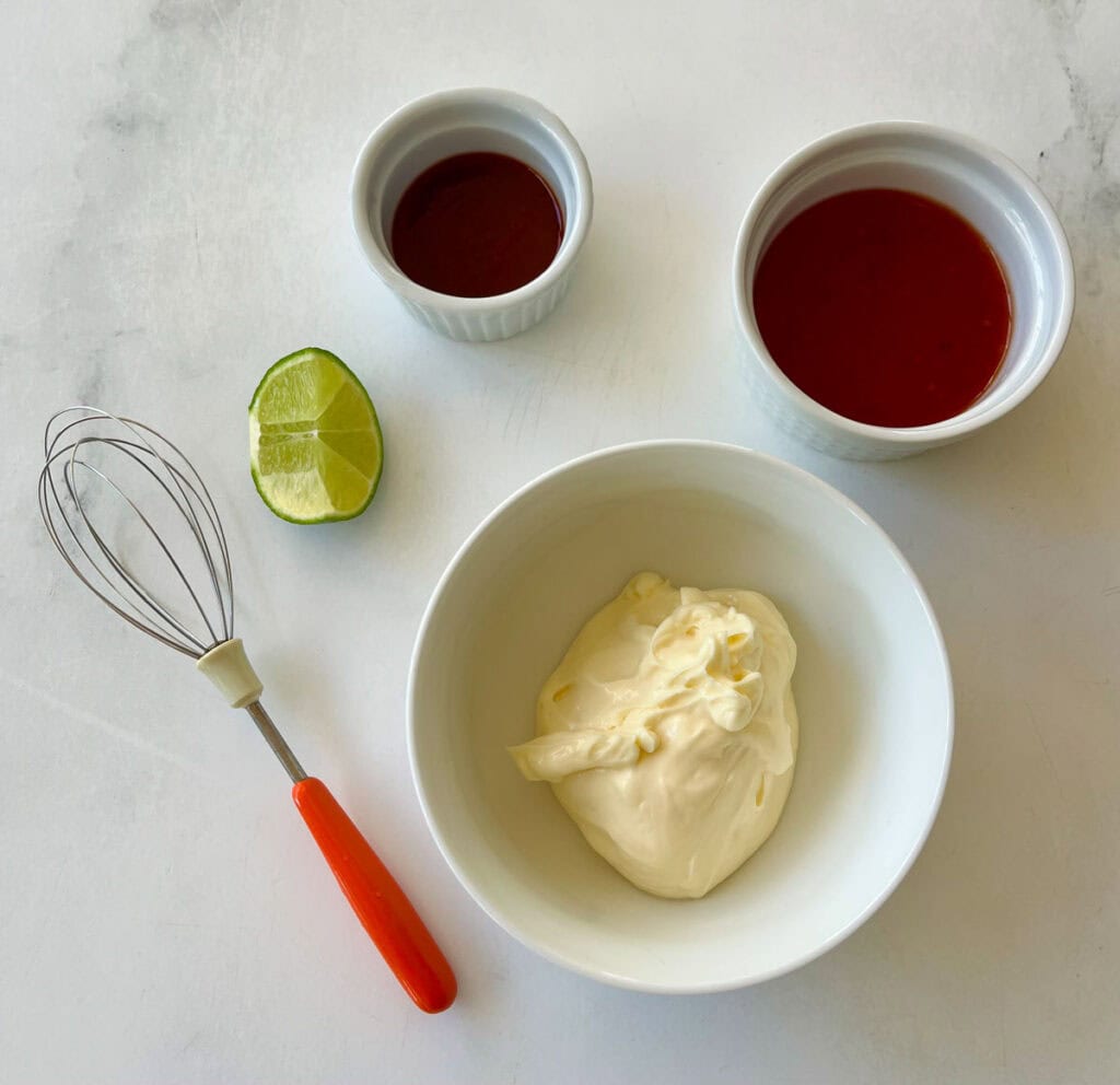 Bowls of mayonnaise, chili sauce, hot sauce, and a wedge of lime. There is a whisk. Items are on a marbled surface.