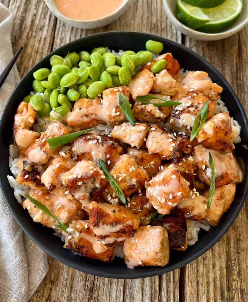 A close-up of seared salmon bowl featuring tender salmon pieces, rice, edamame, and a side of lime wedges and spicy sauce on a rustic wooden table.
