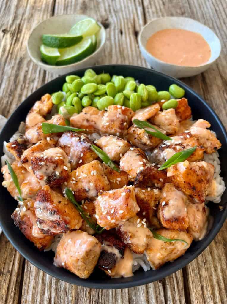 A close-up of seared salmon bowl featuring tender salmon pieces, rice, edamame, and a side of lime wedges and spicy sauce on a rustic wooden table.