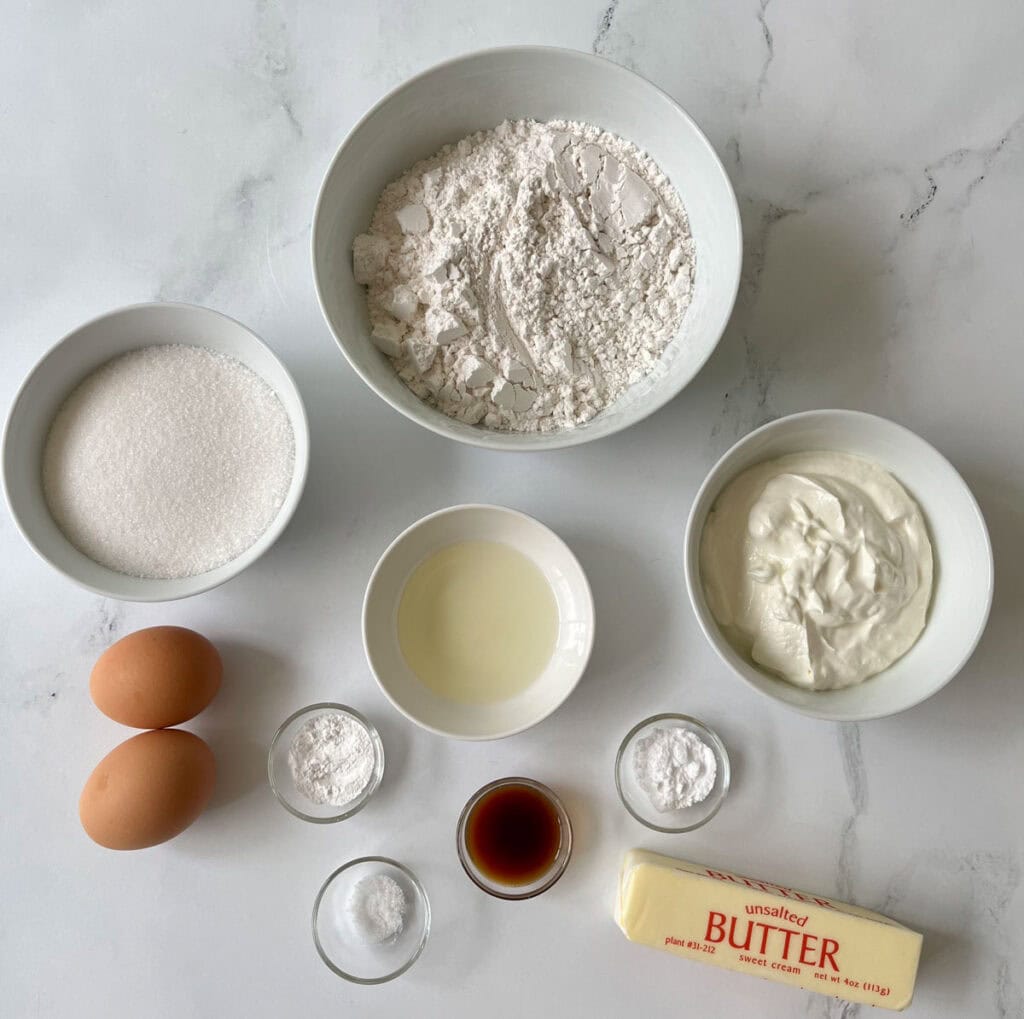 Various baking ingredients such as flour, eggs, butter, sugar, vanilla, and baking powder laid out on a white marble surface.