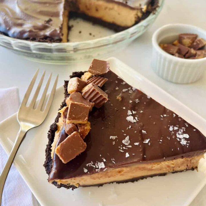 Delicious peanut butter pie slice topped with chocolate pieces, served on a white plate with a fork. There is a whole pie in the background.