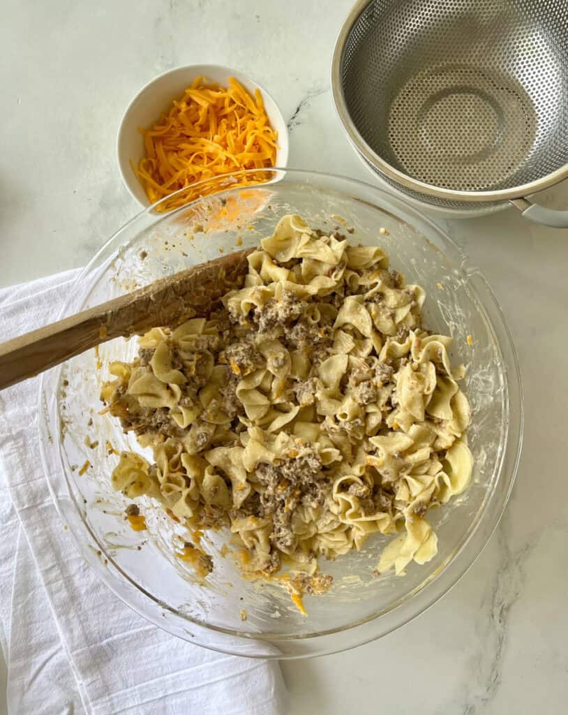 A bowl of cooked ground beef with shredded cheese, fresh noodles, and condensed soup on a white surface.