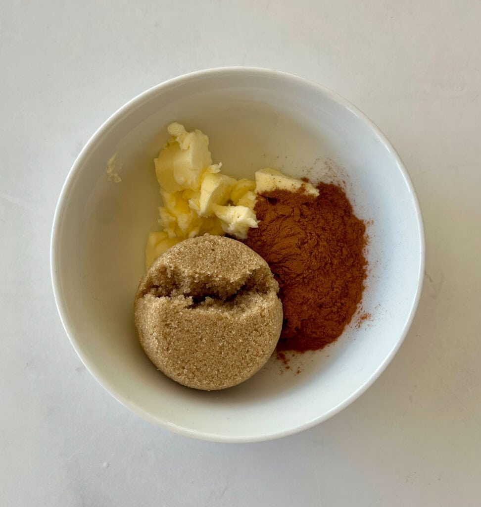 Brown sugar, cinnamon, and butter in a small bowl on a marbled surface.