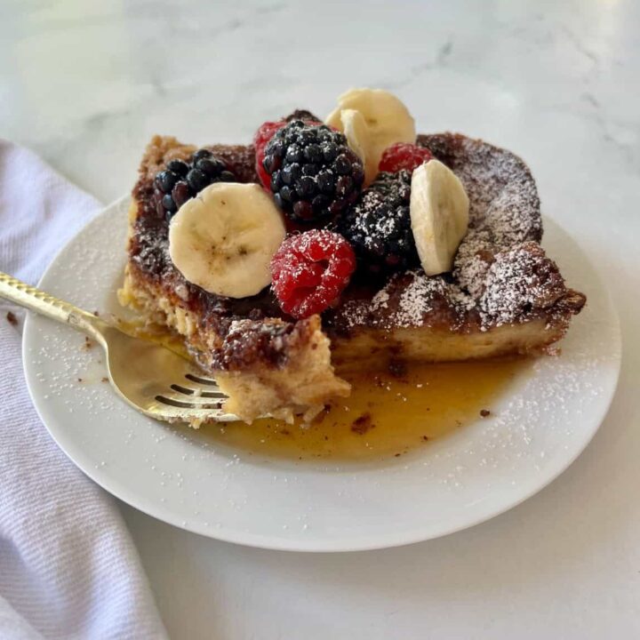 A close-up of a French toast bake topped with mixed berries, banana slices, powdered sugar, and syrup, served on a white plate.