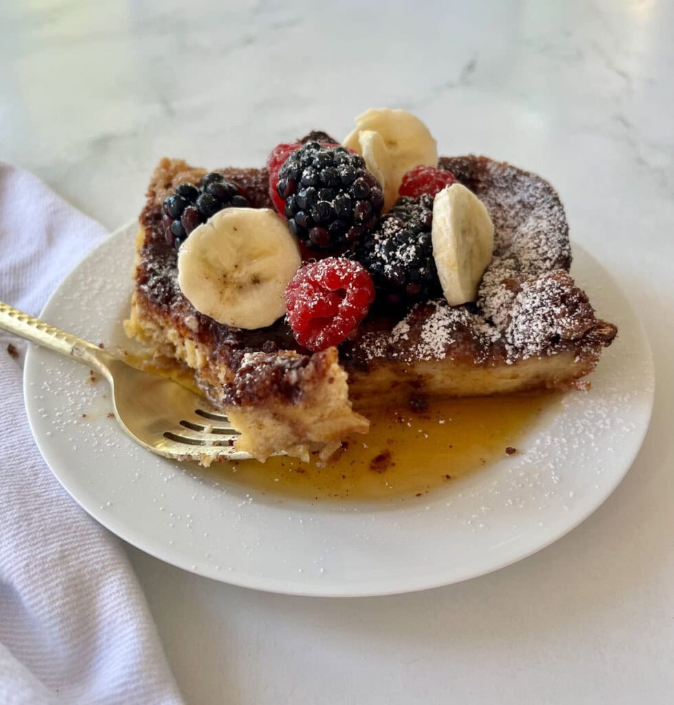 A close-up of a French toast bake topped with mixed berries, banana slices, powdered sugar, and syrup, served on a white plate.