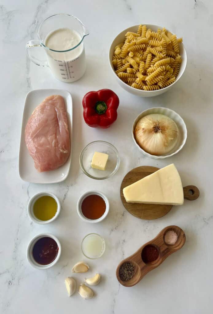 The ingredients for making a delicious creamy chipotle chicken pasta dish, including chicken breast, fusilli pasta, garlic, onion, bell pepper, and seasonings on a white marbled surface.