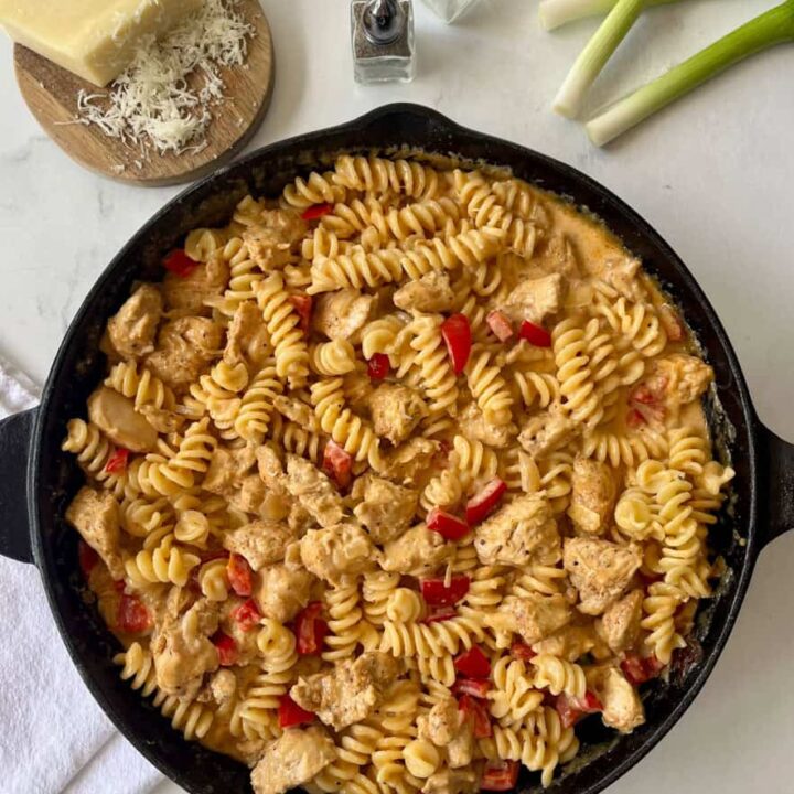 A skillet filled with cooked fusilli pasta, chicken pieces, and red peppers on a marbled surface.