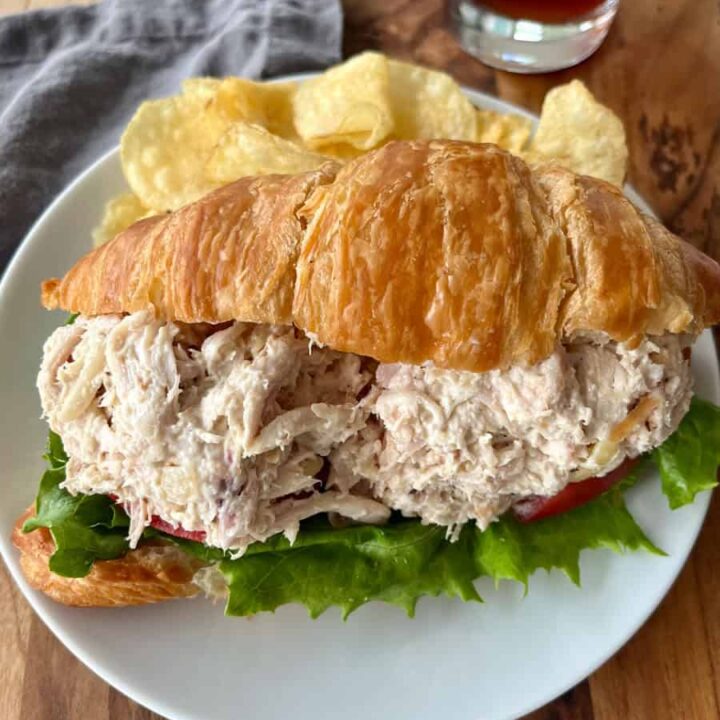 A close-up of a chicken salad croissant sandwich on a white plate, accompanied by potato chips and a glass of iced tea on a wooden table.