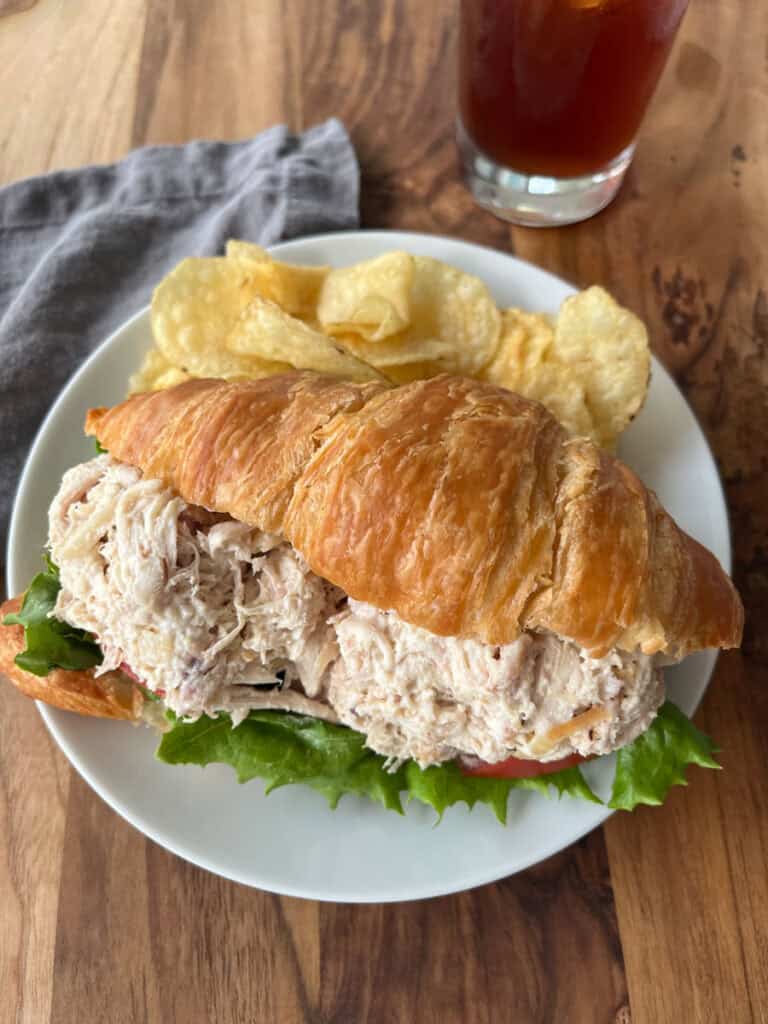 A close-up of a chicken salad croissant sandwich on a white plate, accompanied by potato chips and a glass of iced tea on a wooden table.