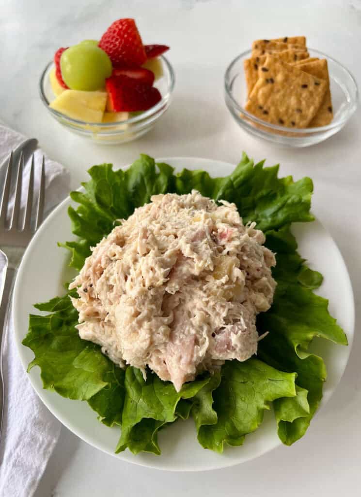 A plate of chicken salad placed on a bed of crisp lettuce, with a side of fresh fruit and crunchy crackers.