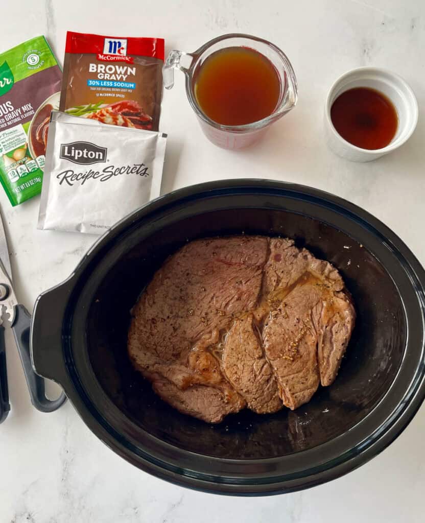 A slow cooker filled with a seasoned beef roast, surrounded by broth, seasoning packets, and spices, ready to cook.