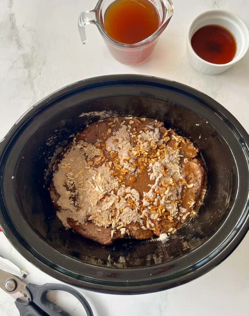 Beef and dry seasonings in a black slow cooker ready to simmer with cups of broth in the background.