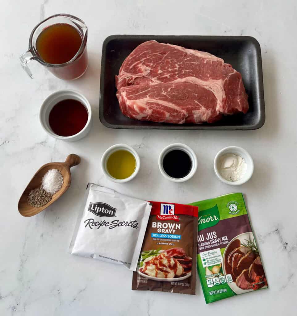 Ingredients for a beef Manhattan, including seasoned beef, broth, gravy mix, and spices on a white surface.