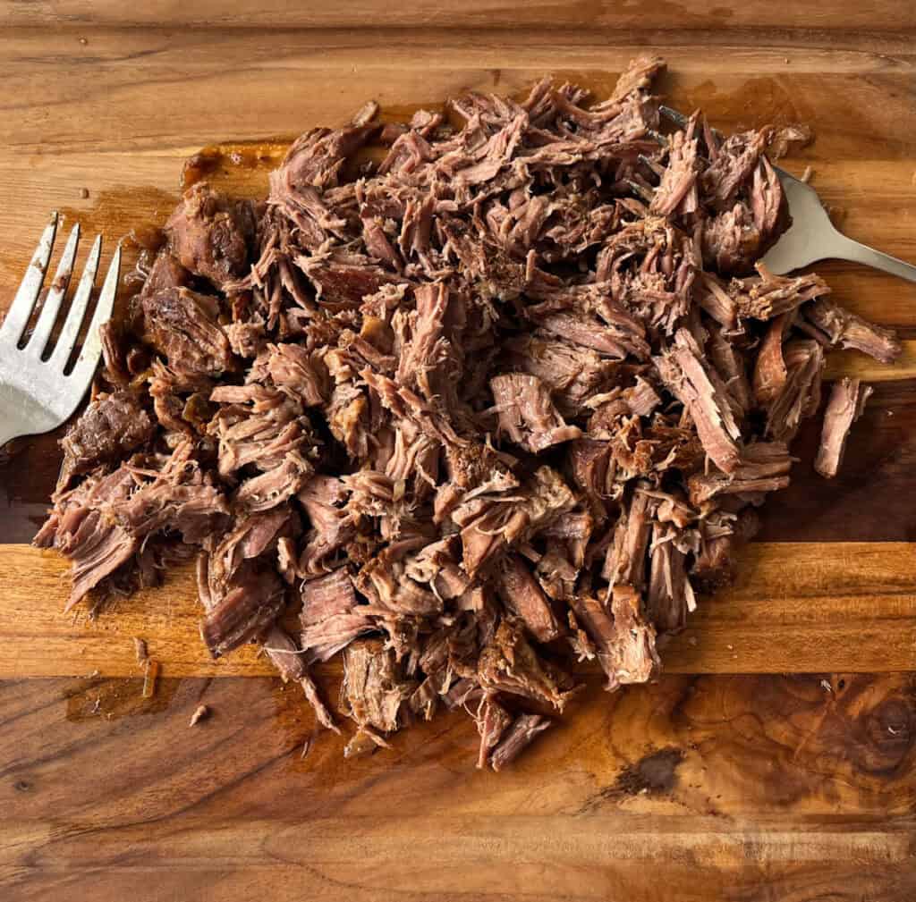 Shredded beef on a wooden cutting board with two large forks.