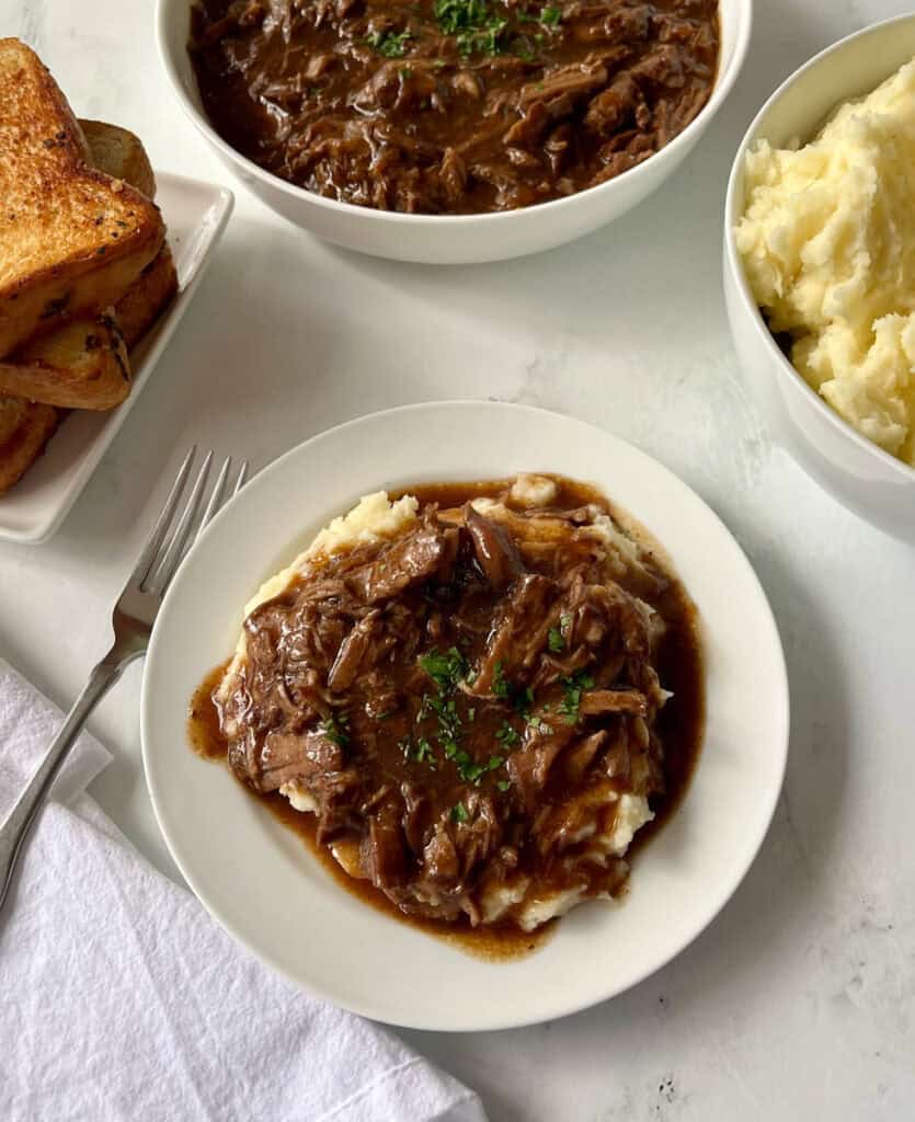 Shredded beef and gravy served over creamy mashed potatoes on a white plate with bowls of food in the background.