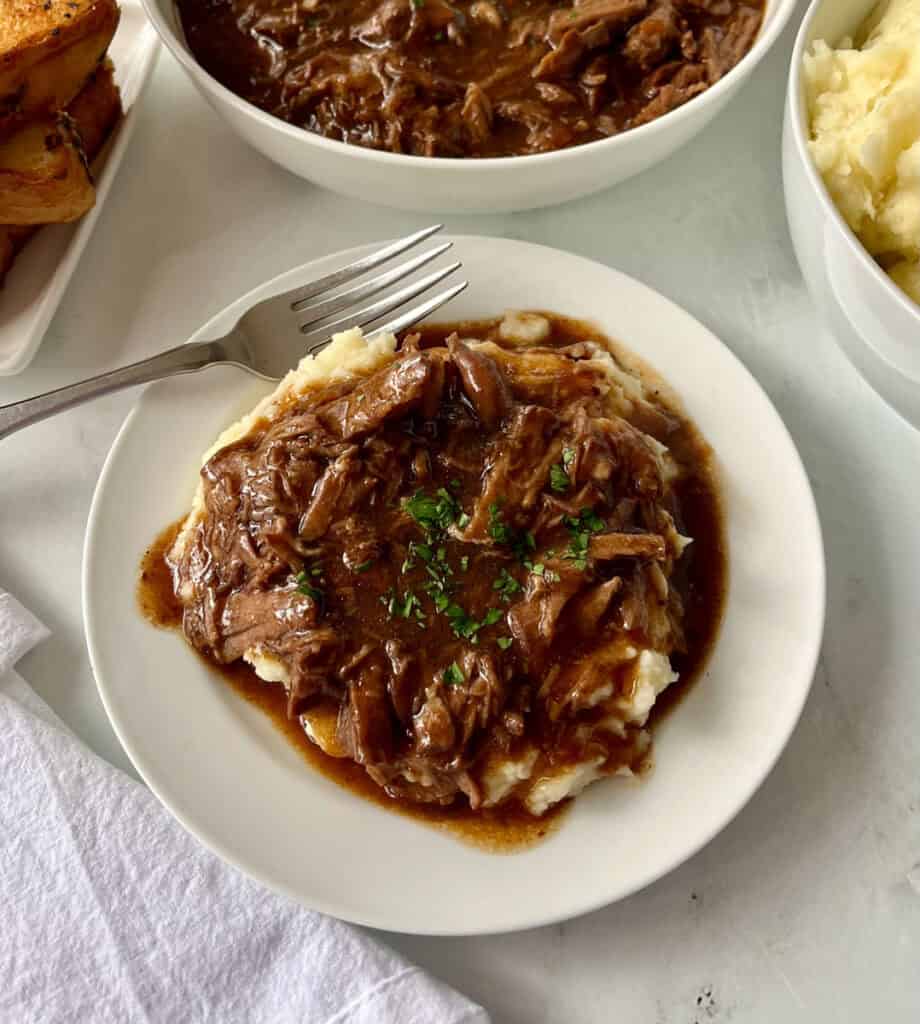 Shredded beef and gravy served over creamy mashed potatoes on a white plate with bowls of food in the background.