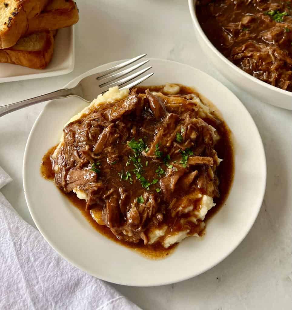 Shredded beef and gravy served over creamy mashed potatoes on a white plate with bowls of food in the background.