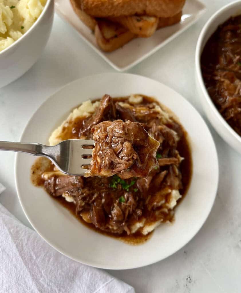 Closeup of shredded beef and gravy served over creamy mashed potatoes on a white plate with bowls of food in the background.