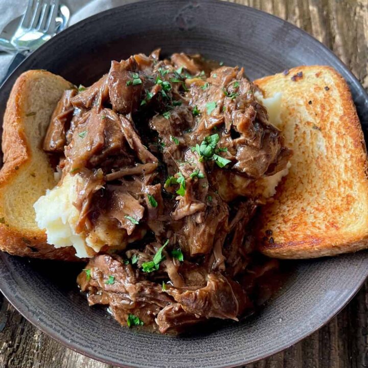 Hearty shredded beef and gravy served over mashed potatoes with toasted bread in a brown bowl on a rustic wooden table.